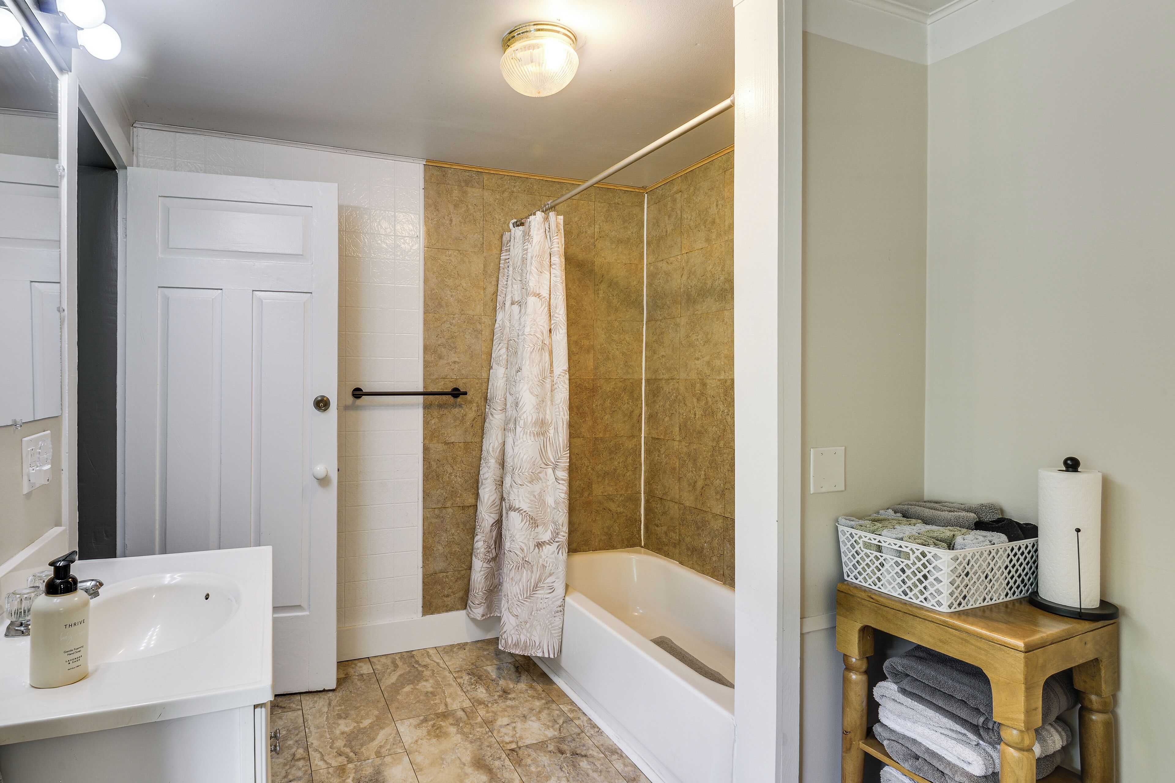 A bathroom with a white sink, a shower curtain, a bathtub, and a wooden side table with towels.