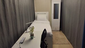 A compact hotel room with a single bed, a desk with a chair, a bedside table, a small potted plant, a wardrobe, and a modern ceiling light fixture.
