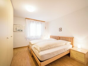 Ein Schlafzimmer mit einem hölzernen Bett, einem Nachttisch mit Lampe, einem Fenster mit transparenten Vorhängen und einem gerahmten Bild an der Wand.