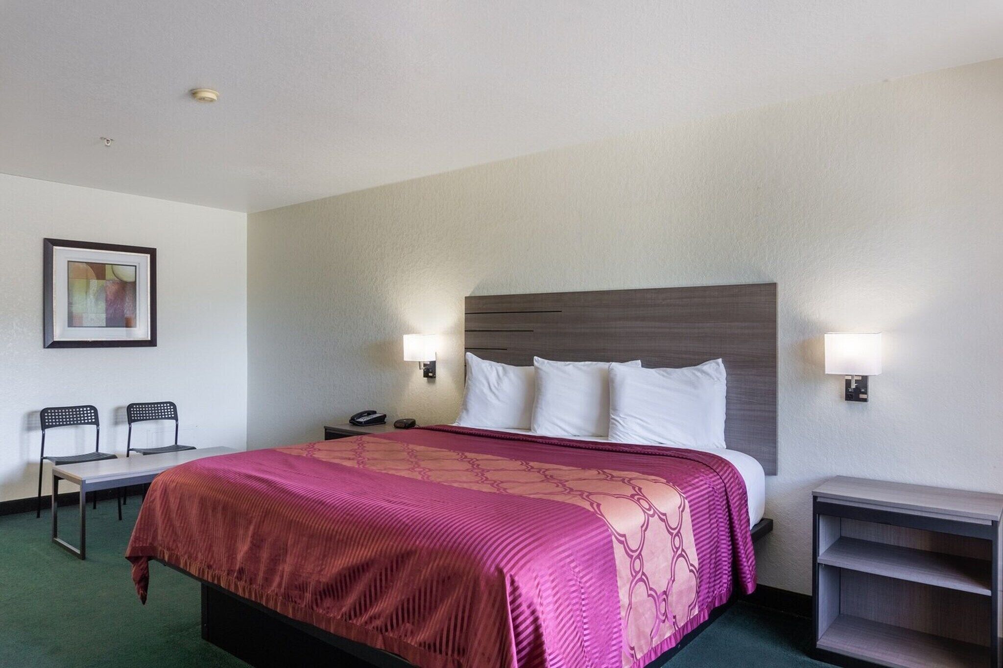 Une chambre d’hôtel avec un lit, des tables de chevet, un bureau, deux chaises et un tableau encadré au mur.