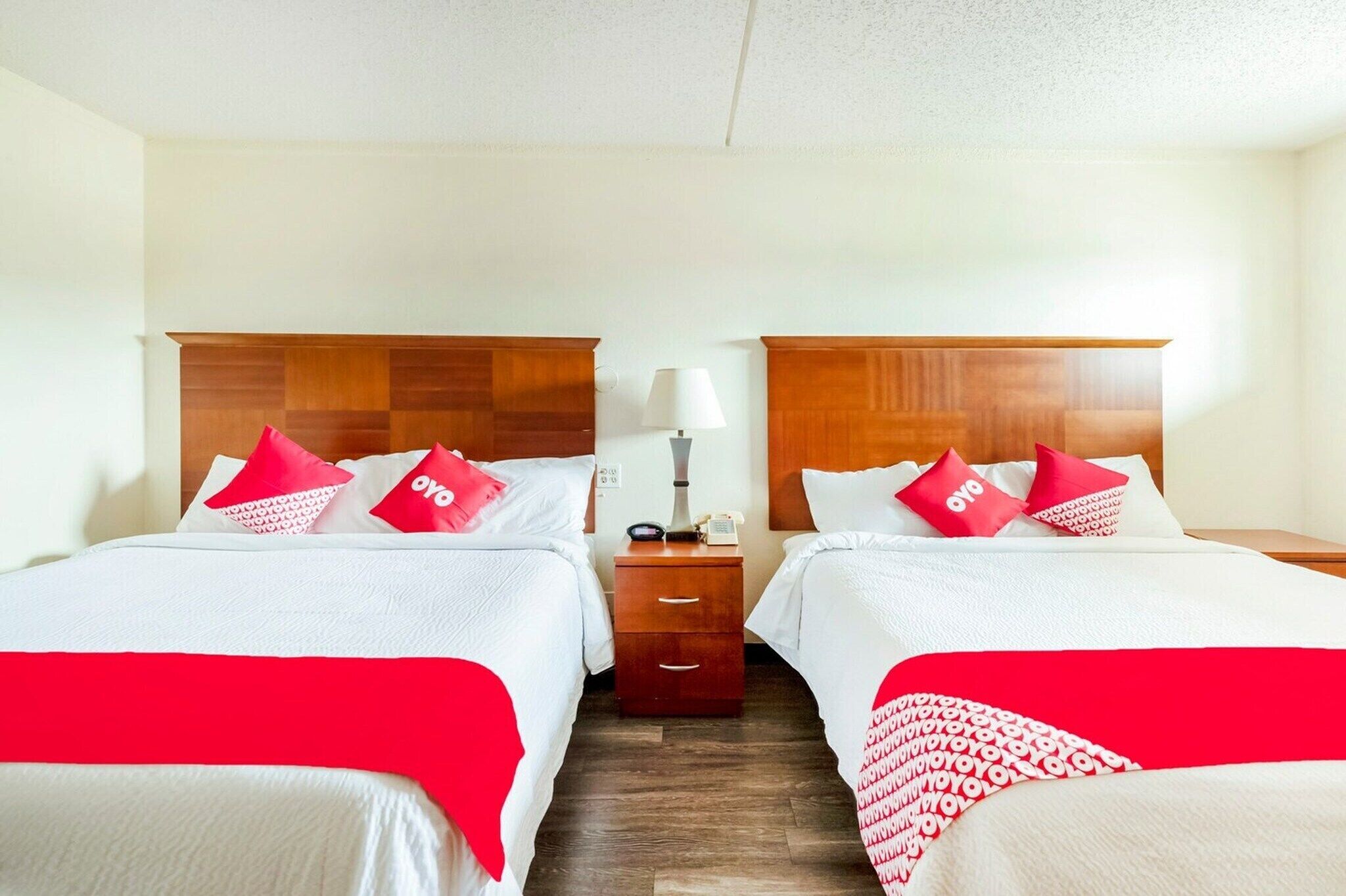 Ein Hotelzimmer mit zwei Betten, jeweils mit Holz Kopfteil, weißer Bettwäsche und roten Kissen mit 'oyo'-Branding. Ein Nachttisch mit Lampe und Telefon ist vorhanden.