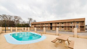 Uma piscina com deck de madeira, mesa de piquenique e um edifício com varandas ao fundo.