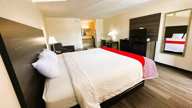 Quarto de hotel com cama, escrivaninha, cadeira, televisão, espelho e banheiro ao fundo.