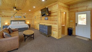 Interior de una cabaña de madera con cama, sofá, televisor y ventilador de techo.