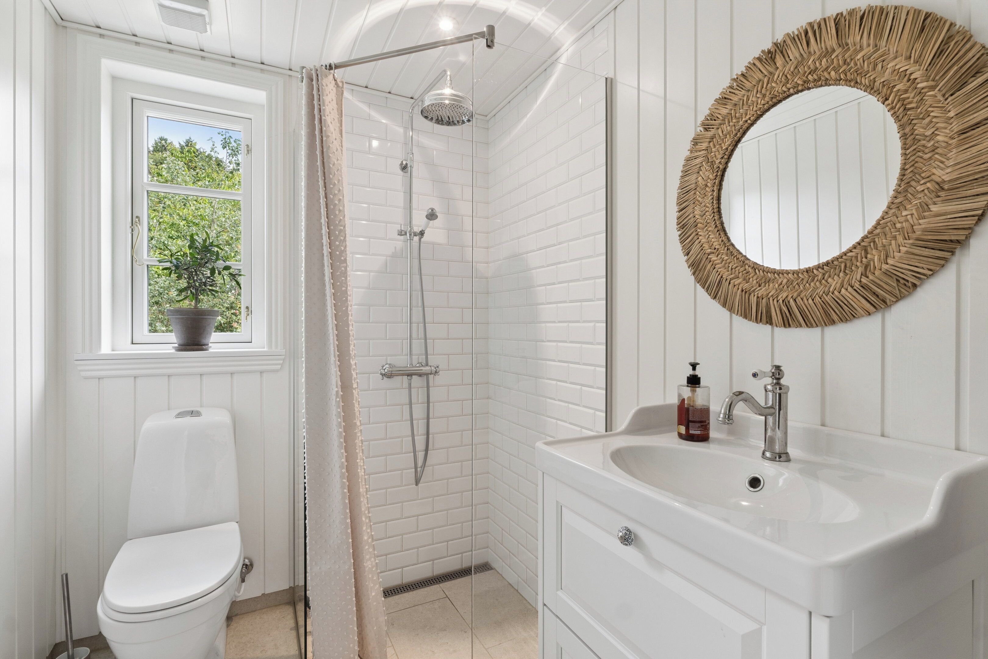 Ein Badezimmer mit Toilette, einer Dusche mit Glastür, Waschbecken mit Spiegel und einem Fenster mit Blumentopf.