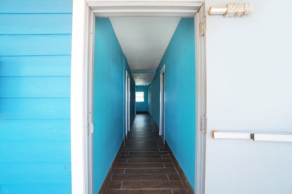 A hallway with blue walls and a dark floor, leading to a door with a light switch and a wall-mounted light fixture. - Corpus Christi Tx | Queen Bed (Corpus Christi TX)