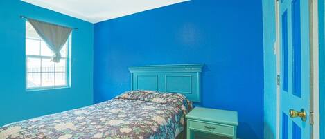 Um quarto com cama, mesa de cabeceira, janela com cortina e uma porta azul.