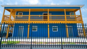 Un bâtiment à deux étages, avec une façade bleue et jaune, un balcon à l’étage supérieur et une clôture en métal noir au premier plan.
