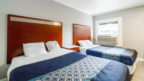 A hotel room with two beds, a wooden headboard, a window with an air conditioning unit, and a white wall.