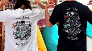 Una habitación con una pared cubierta de textiles estampados, una cama con almohadones coloridos, y dos personas con camisetas con texto.