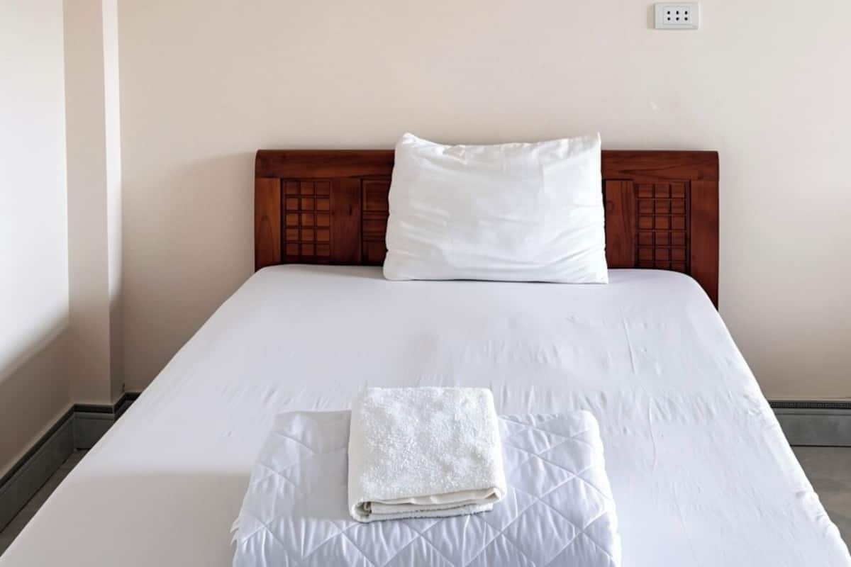A neatly made bed with a folded white towel and a pillow, a wooden headboard, and a white wall with a power outlet.