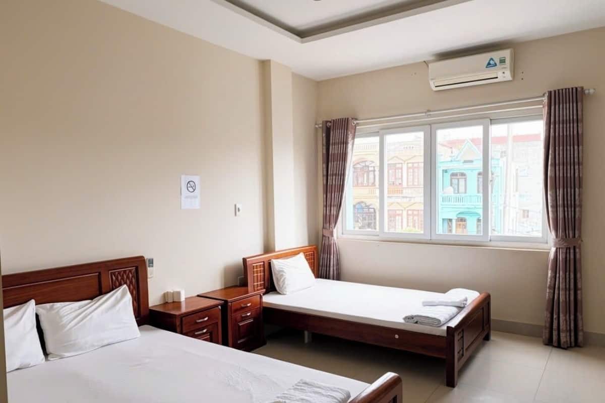 A hotel room with two beds, a window with curtains, an air conditioning unit, and a tiled floor.