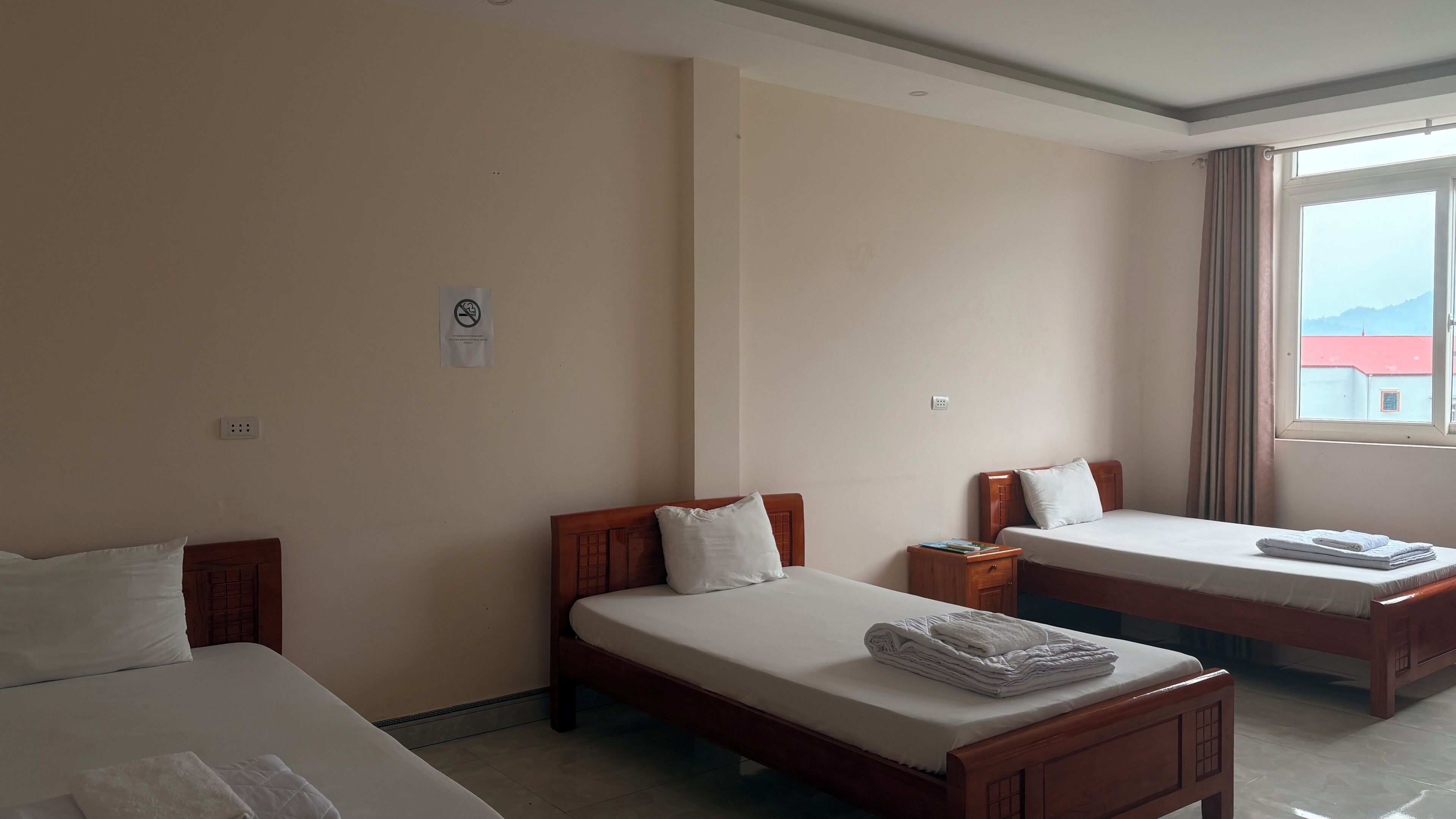 Habitación de hotel con dos camas individuales, una mesita de noche de madera, una ventana con cortinas y vistas a edificios.