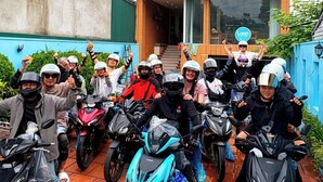 Un grupo de personas con cascos y motocicletas frente a un edificio azul con unidades de aire acondicionado.