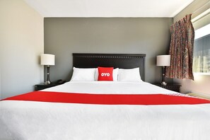 A hotel room with a large bed, two bedside tables with lamps, a red throw blanket, and a window with curtains. - Queen Bed | Non smoking (Shreveport LA)