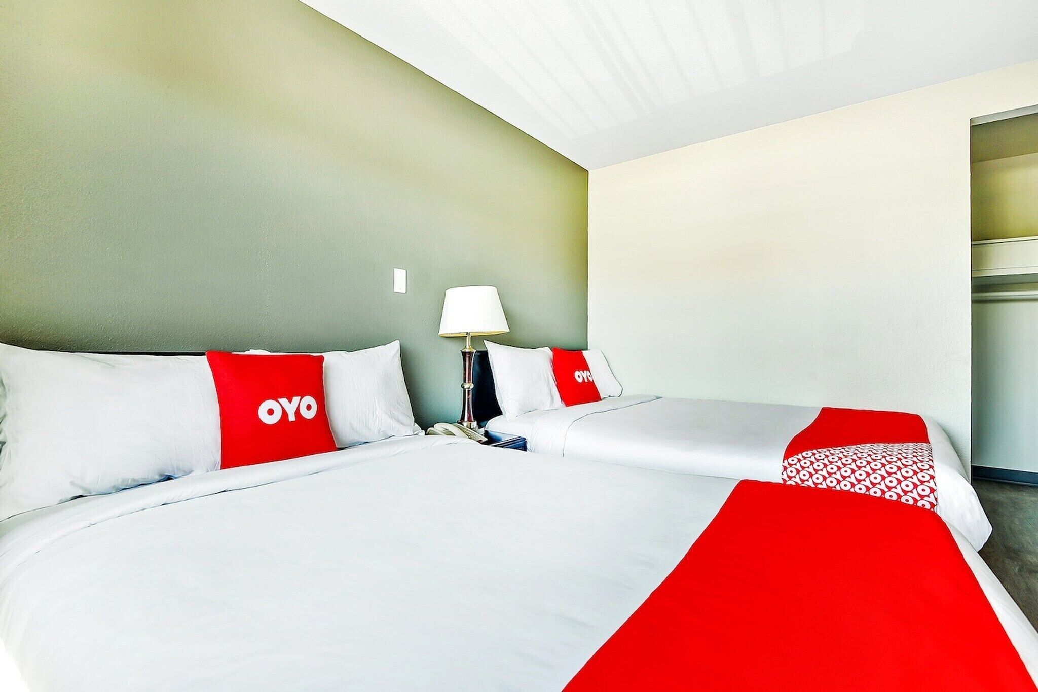 Habitación de hotel con dos camas, cada una con ropa blanca y detalles rojos, una mesita de noche con lámpara y una pared de color neutro.