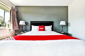 A hotel room with a bed, two bedside lamps, a window with curtains, and a red pillow with 'OYO' on it. - Premium King Bed Smoking (Shreveport LA)