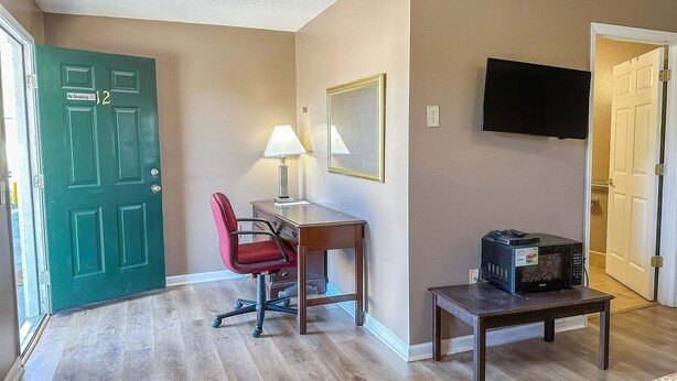 A hotel room with a green door, a desk with a lamp, a television, and a microwave on a small table. - Queen Bed | Smoking | Sparta, GA (Sparta, GA)