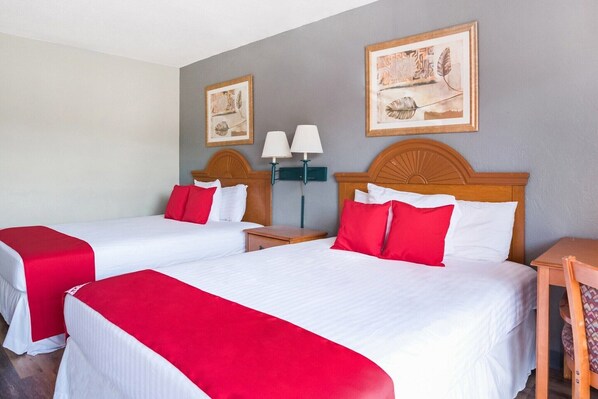 A hotel room with two beds, each with a red bedspread and white linens, a wooden headboard, bedside tables, and wall-mounted lamps.