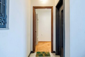 Un couloir étroit, avec une porte en bois au fond, un petit tapis sur le sol et une fenêtre avec une grille en métal sur le côté gauche.