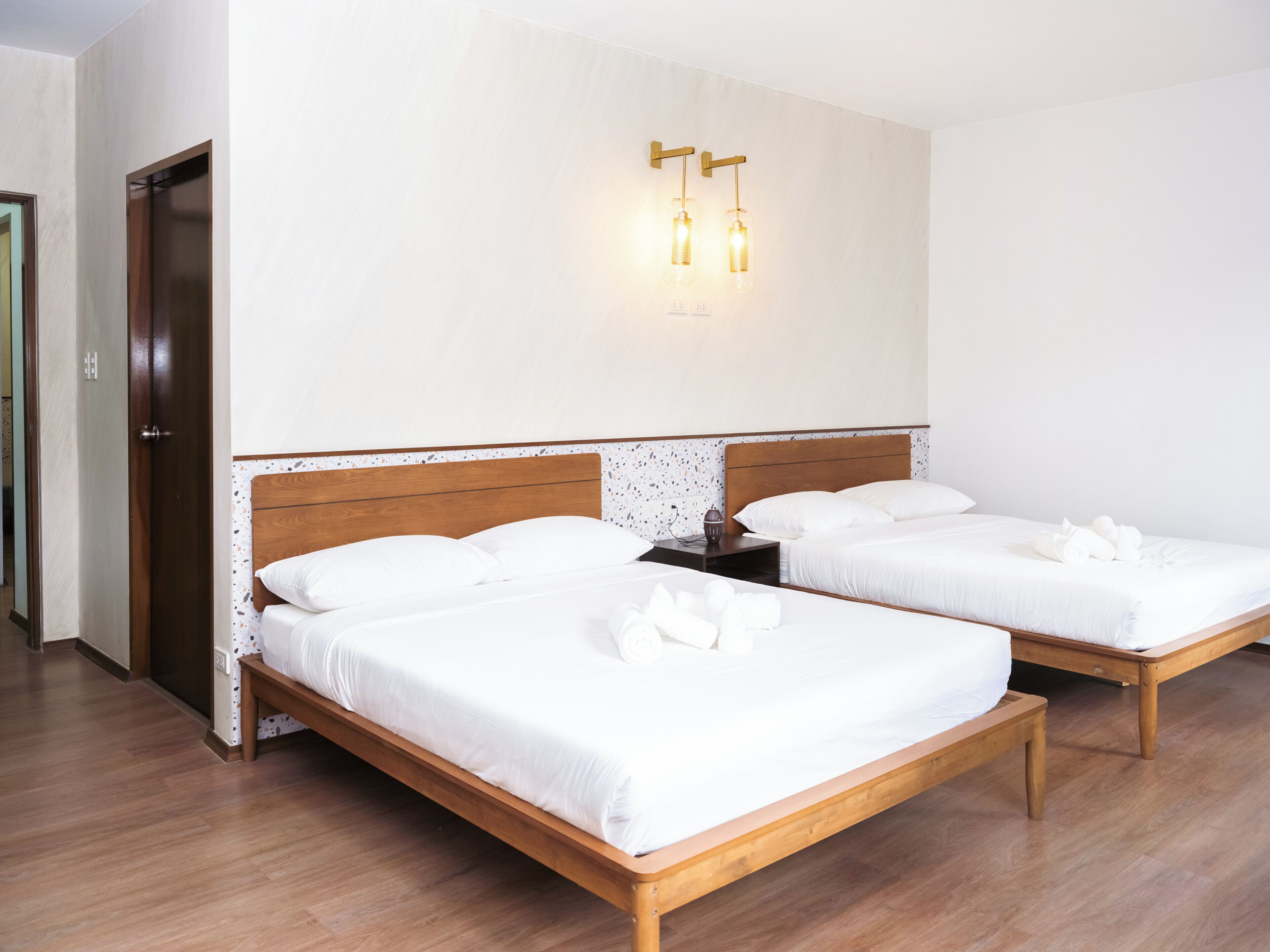 Une chambre d’hôtel avec deux lits en bois, une table de chevet en bois, un luminaire fixé au mur et une porte.
