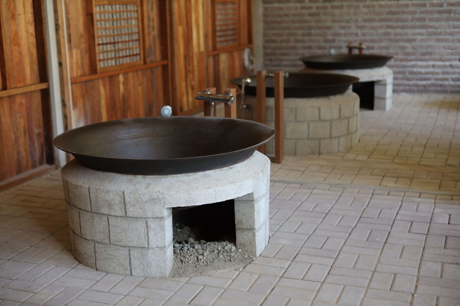A traditional cooking area with a large wok on a stone stove, a brick wall, and a tiled floor.