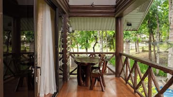 Una terraza de madera con una mesa, sillas, un ventanal y vistas a la vegetación.