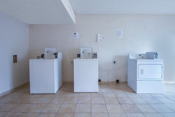 A laundry room with three white appliances, a wall-mounted thermostat, and a small trash bin.