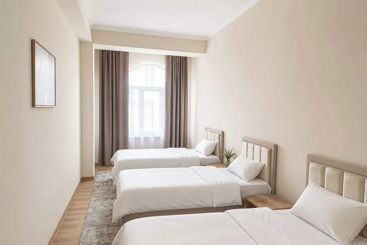A hotel room with three beds, a large window with curtains, a wooden floor, and a ceiling light.