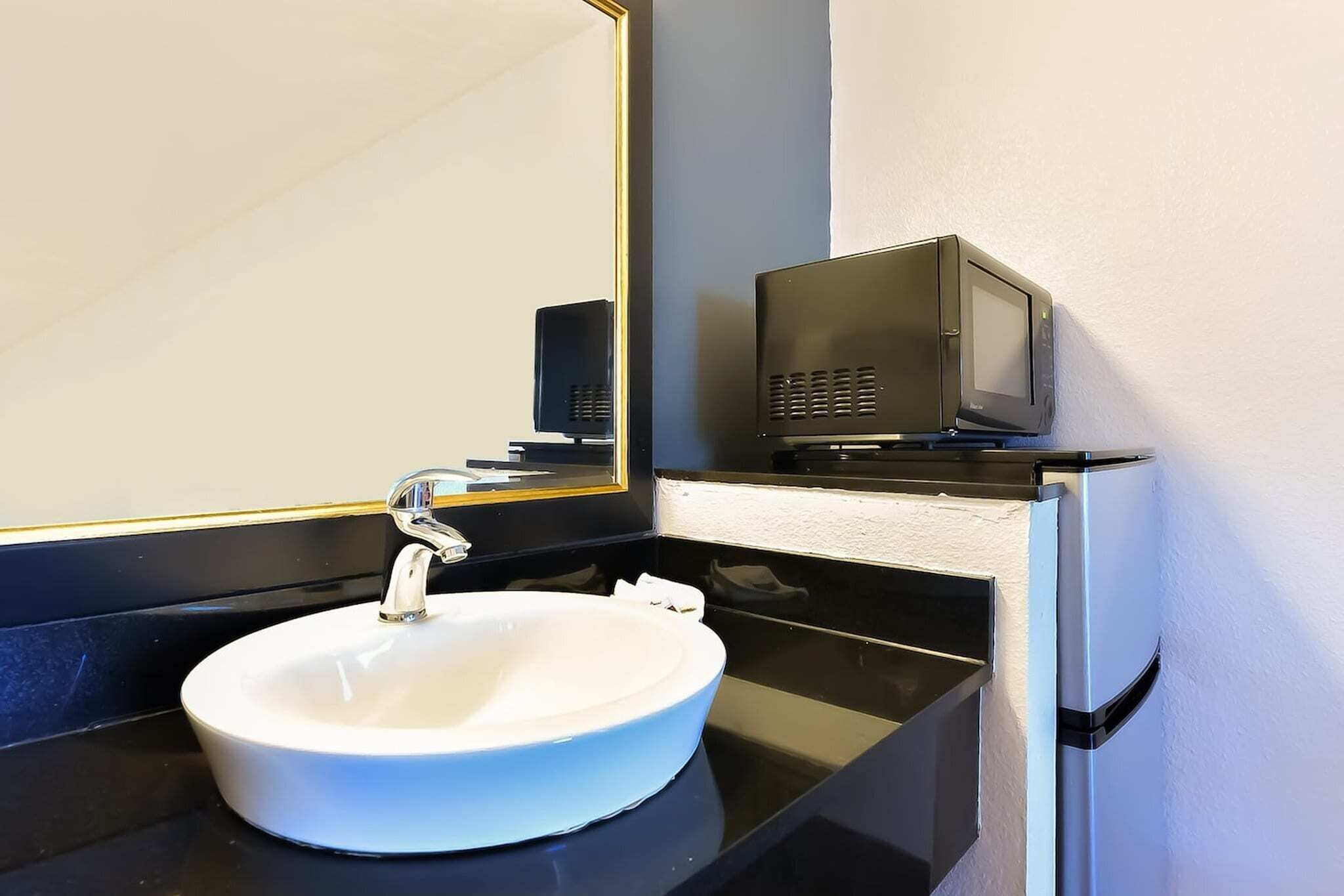 Un bagno con un lavandino bianco, uno specchio, un piano di lavoro nero e una televisione su una piccola mensola.