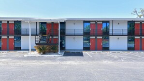 Un motel de varios pisos con una escalera central, estacionamiento y una pequeña zona ajardinada con plantas.