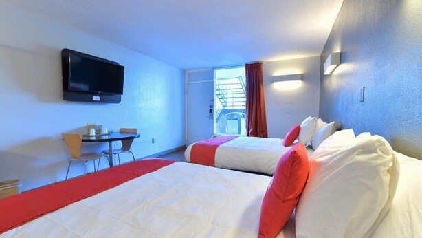 Une chambre d’hôtel avec deux lits, un téléviseur fixé au mur, une petite table avec des chaises et une fenêtre avec des rideaux.