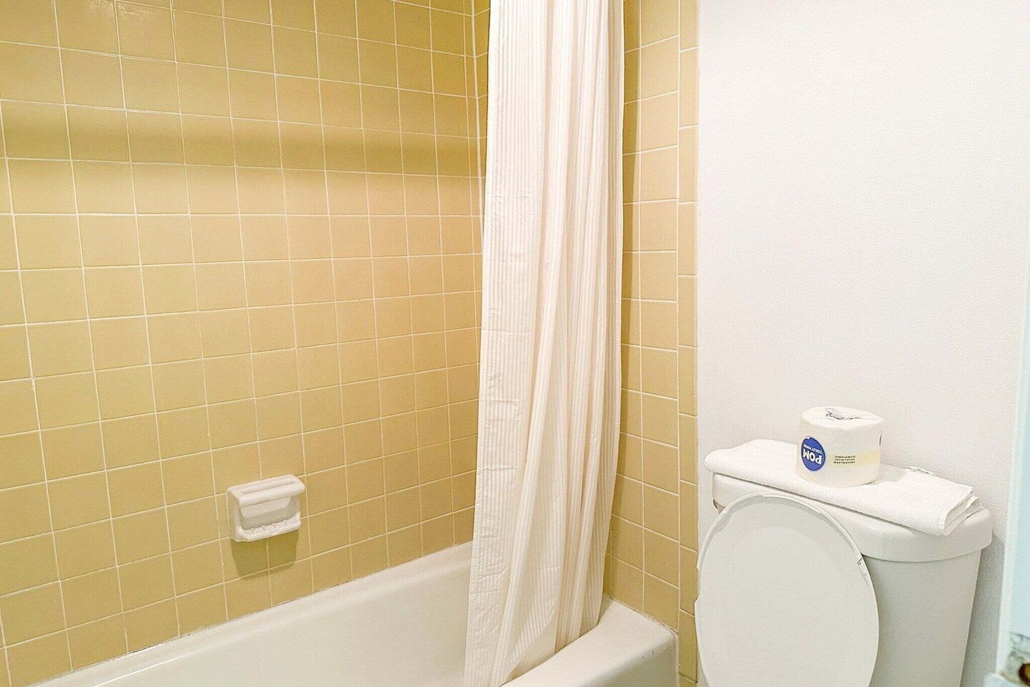 Un bagno con pareti piastrellate di giallo, una vasca da bagno bianca, un water bianco con porta carta igienica e un telo da doccia bianco.