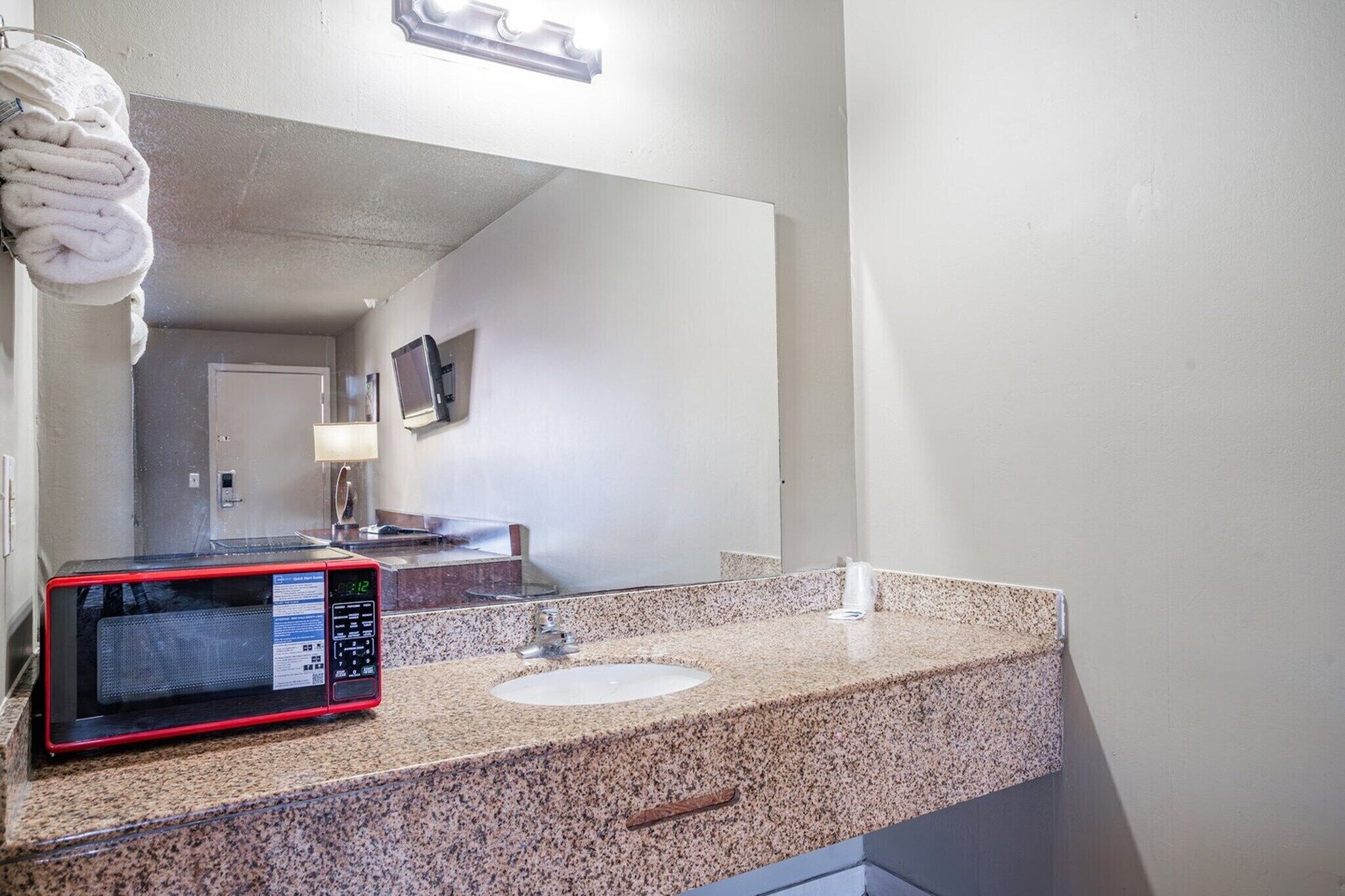 A bathroom with a granite countertop, a sink, a mirror, a towel rack, a microwave, and a television.