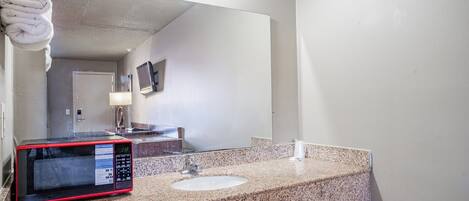 A bathroom with a granite countertop, a sink, a mirror, a towel rack, a microwave, and a television.