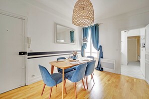 A dining area with a wooden table, blue chairs, a hanging light fixture, a mirror, and a window with curtains. - Comfortable Apartment 1BR/4P - Champs Élysées (Paris)