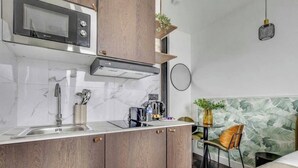 Een compacte keuken met een magnetron, spoelbak en houten kastjes. Er is een klein eethoekje met een stoel en tafel, en een sierplant.