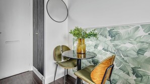 Een kamer met een ronde tafel, twee stoelen, een bloempot, een spiegel aan de muur en een deur.