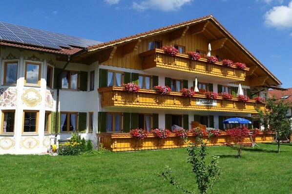 Ein zweistöckiges Hotel mit einem mit Blumen geschmückten Balkon, einem Schild mit der Aufschrift 'Hotel Alpenblick' und einer Solaranlage auf dem Dach.