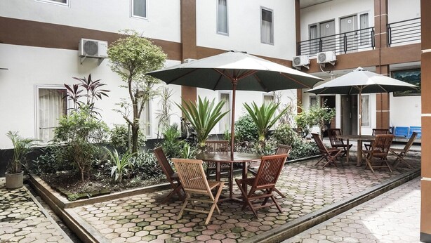 Un patio con mesa y sillas bajo sombrillas, rodeado de plantas en macetas y un suelo de baldosas.
