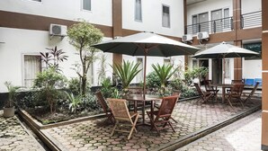 Un patio con mesa y sillas bajo sombrillas, rodeado de plantas en macetas y un suelo de baldosas.