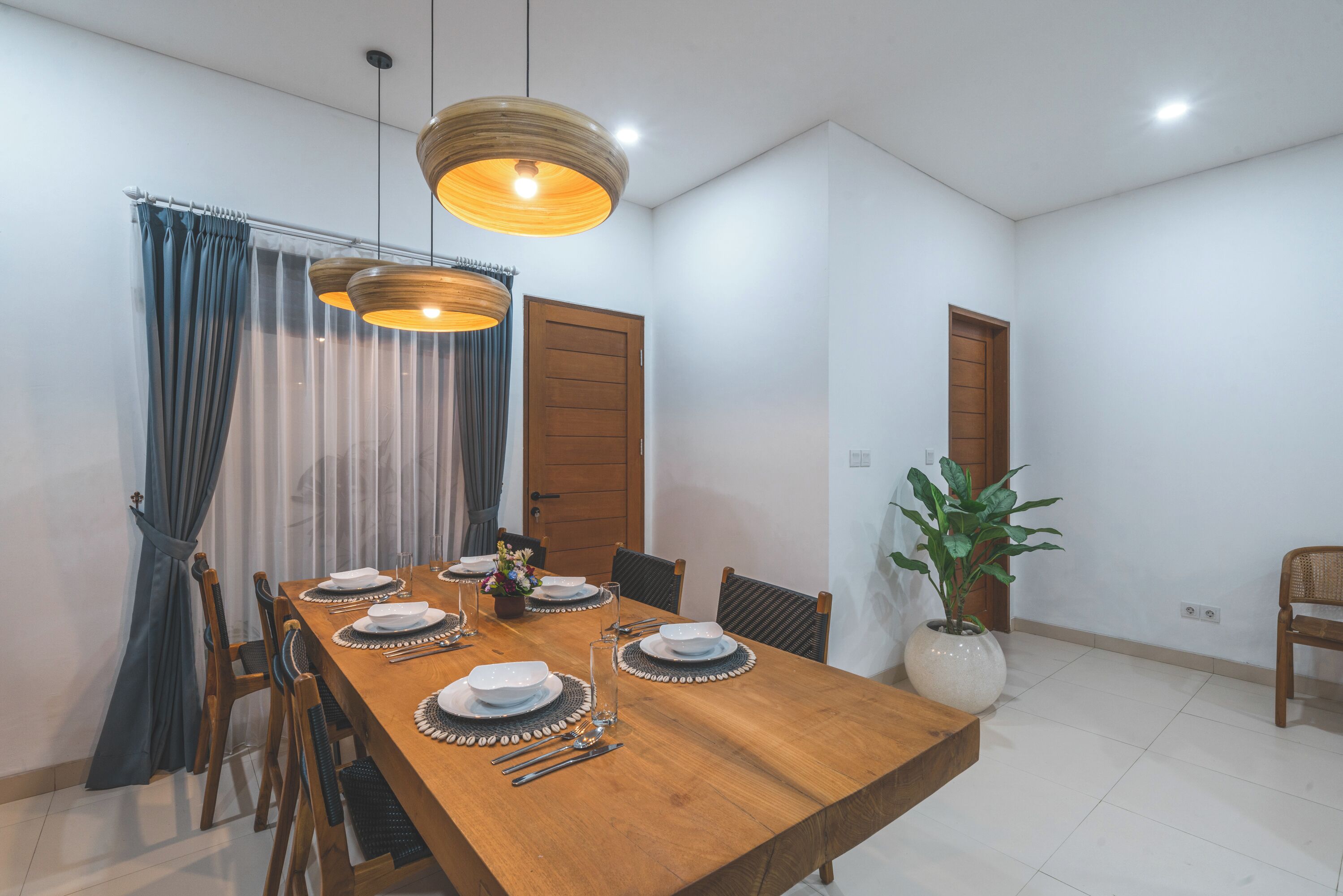 A dining area with a wooden table set for four, two pendant lights, a potted plant, and a door in the background.