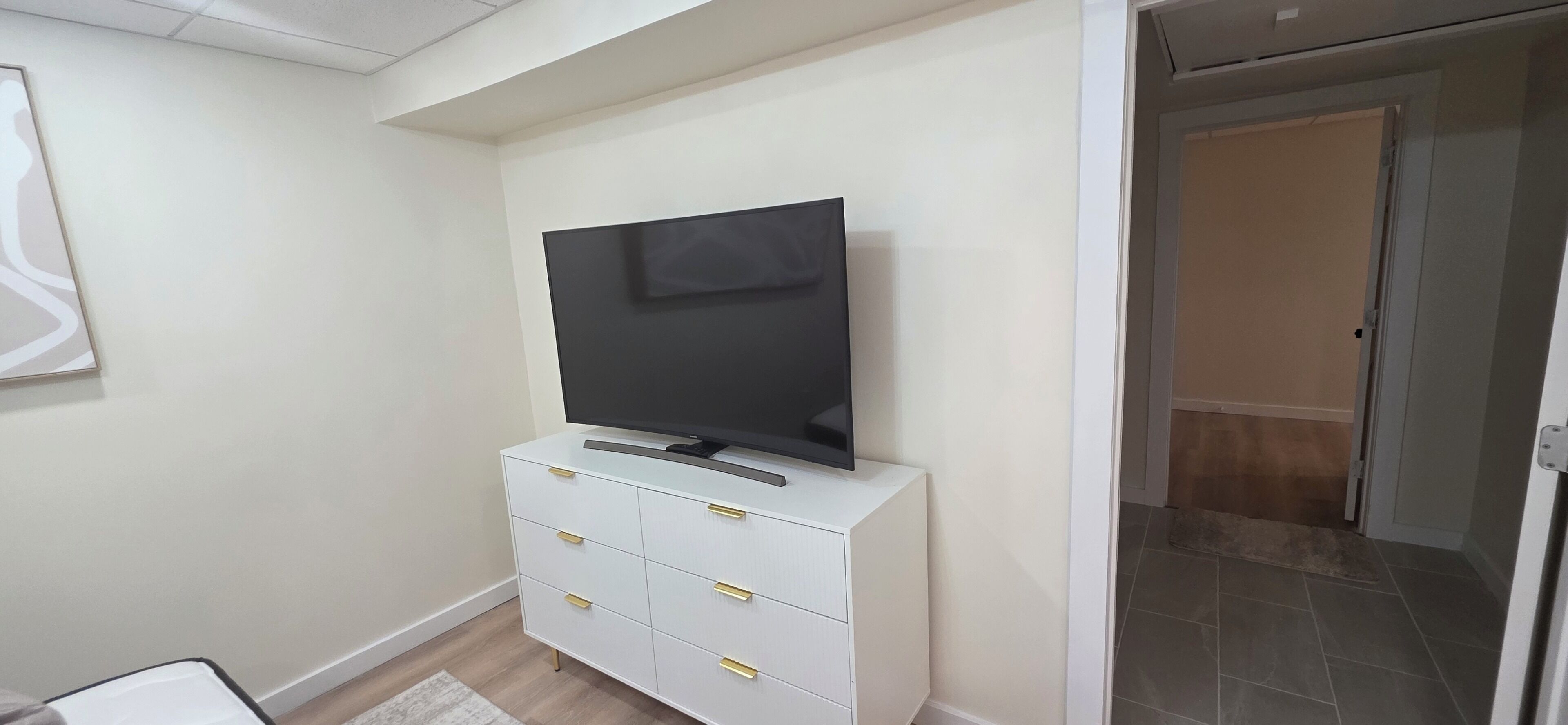 Eine weiße Kommode, ein Flachbildfernseher, ein gerahmtes Bild an der Wand und eine Tür zu einem anderen Zimmer.