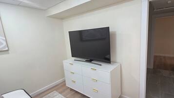 Eine weiße Kommode, ein Flachbildfernseher, ein gerahmtes Bild an der Wand und eine Tür zu einem anderen Zimmer.