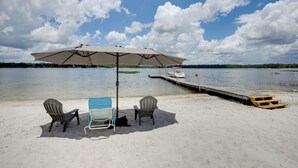 Een strand met een tafel en stoelen onder een parasol, een steiger en een boot op de achtergrond.