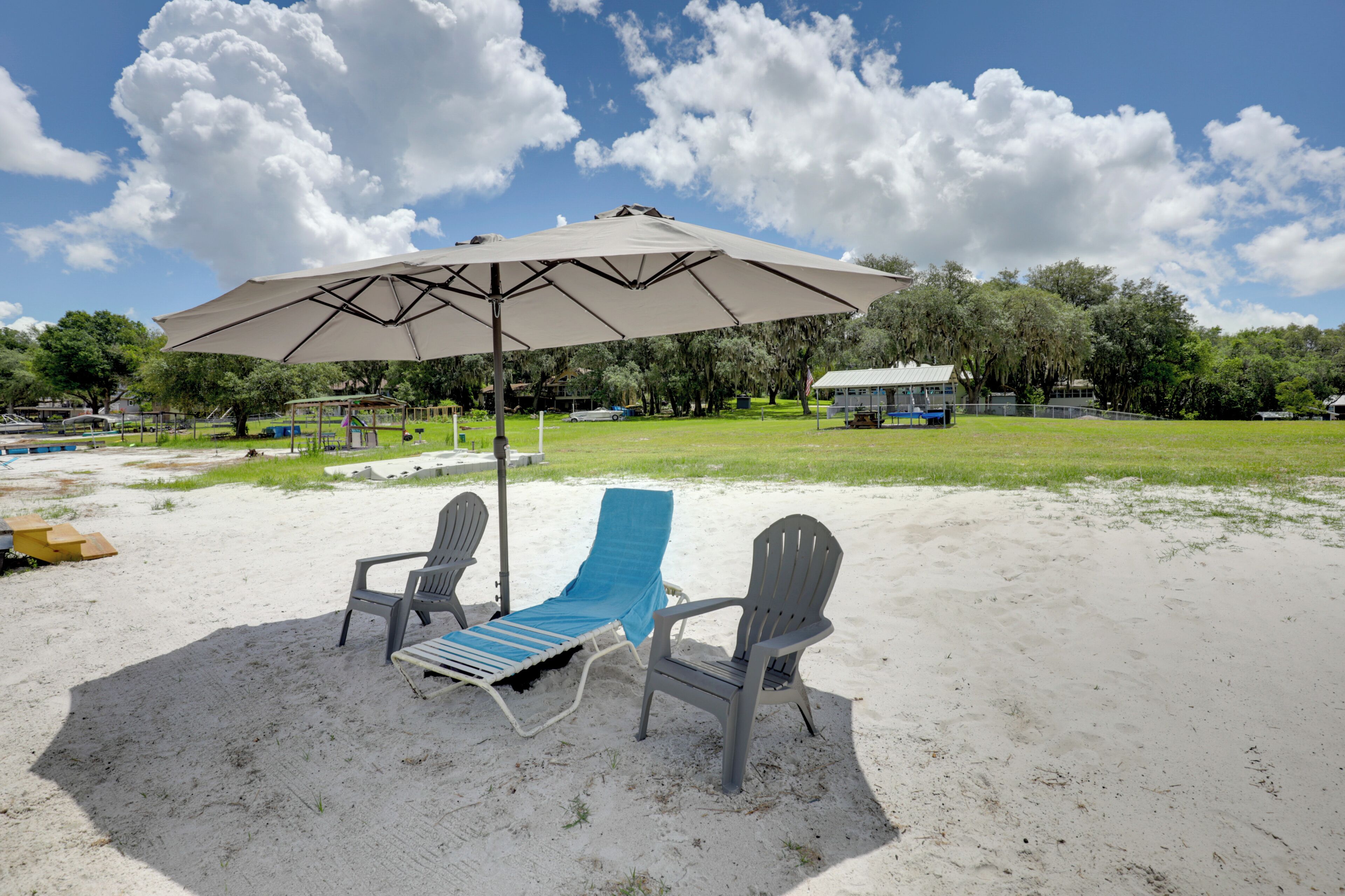A beach with two lounge chairs and a large umbrella, a grassy area, and trees in the background.