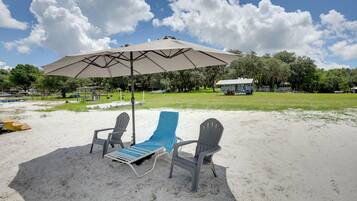 A beach with two lounge chairs and a large umbrella, a grassy area, and trees in the background.