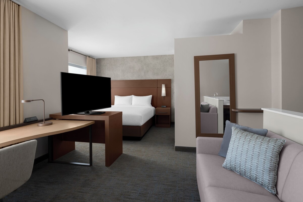 A hotel room with a bed, a desk with a television, a bedside table, a sofa, and a bathroom with a mirror and sink.