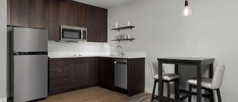 A modern kitchen with dark wood cabinets, a stainless steel refrigerator, a microwave, and a sink. There is a dining area with a table and two chairs.