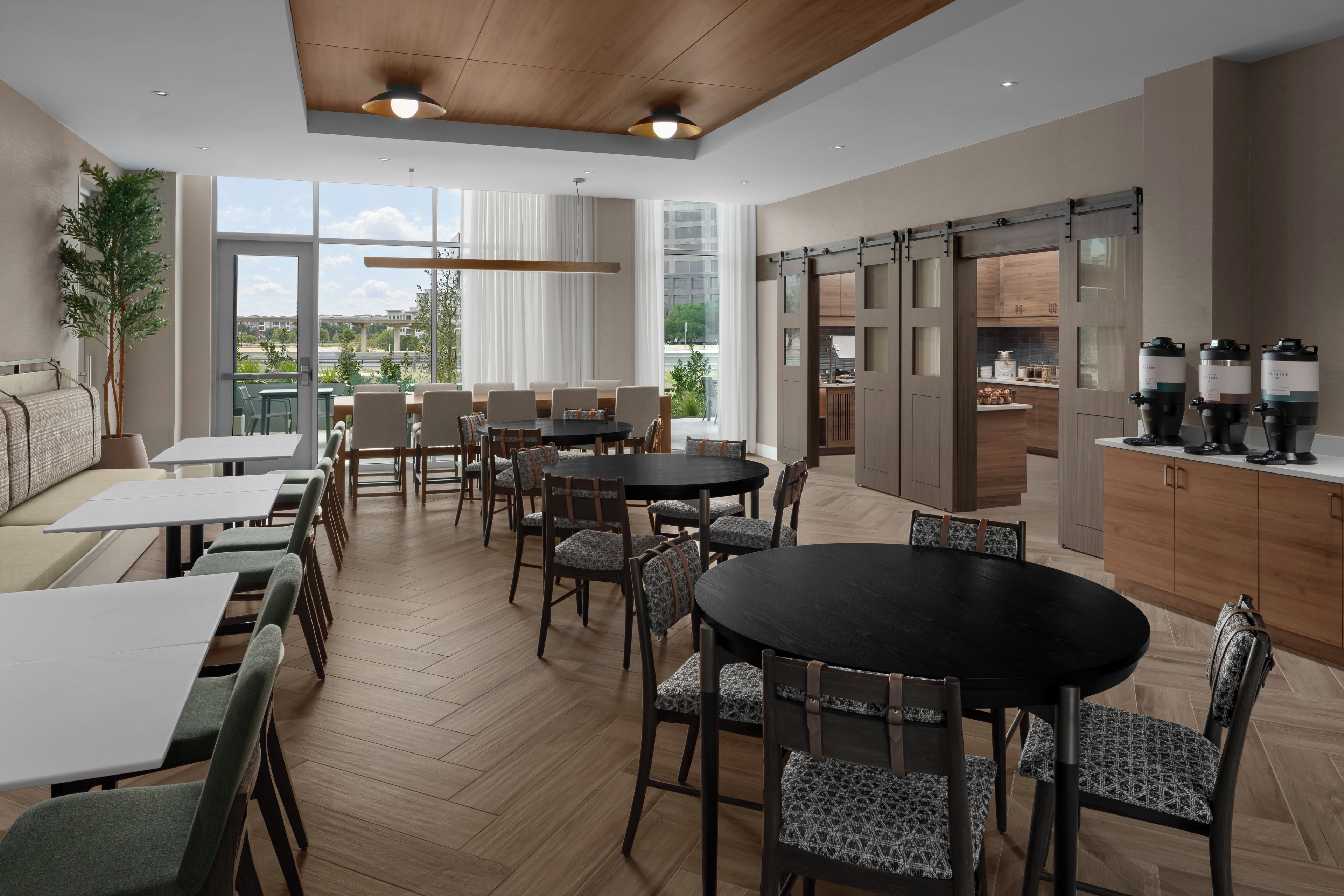 A modern dining area with multiple tables and chairs, large windows offering a view of the outdoors, and a kitchen area with appliances.